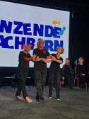Drei ältere Erwachsene in schwarzer Kleidung halten sich an den Händen und tanzen lächelnd auf der Bühne. Hinter ihnen wird auf einem großen Bildschirm ein fetter blauer und gelber Text angezeigt. Andere Menschen sitzen im Hintergrund und beobachten die Aufführung.