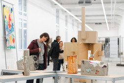 Eine Frau lächelt, während sie die abstrakten Skulpturen in einer hellen, modernen Kunstgalerie betrachtet. Im Hintergrund sind mehrere Menschen zu sehen, und an den Wänden hängen bunte Kunstwerke.