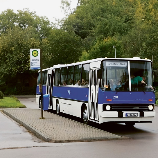 An einem regnerischen Tag steht ein blau-weißer Bus an einer Bushaltestelle. Das angezeigte Ziel lautet „Karl-Marx-Stadt“. Der Bus steht auf einem gepflasterten Platz neben einer nassen Straße, umgeben von üppigen grünen Bäumen.