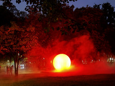 A bright yellow sphere surrounded by red mist illuminates a park at dusk, with trees and the silhouettes of people visible in the background.