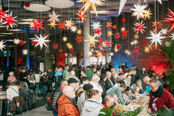 Eine große Menschenmenge versammelt sich drinnen unter vielen hängenden, bunten sternförmigen Laternen. Die Menschen unterhalten sich, essen und nehmen an festlichen Aktivitäten in einem lebhaften, gut beleuchteten Raum teil.