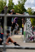 A group of people gather on a graffiti-covered skate park ramp on a cloudy day, some sitting, some standing, with trees and greenery in the background.