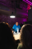 Ein Mann in einem blauen T-Shirt steht hinter einer Theke unter einer Hängelampe in einem schwach beleuchteten Raum mit freiliegenden Ziegelwänden und blickt auf zwei Personen, die der Kamera den Rücken zuwenden. Auf dem Tresen stehen Flaschen.