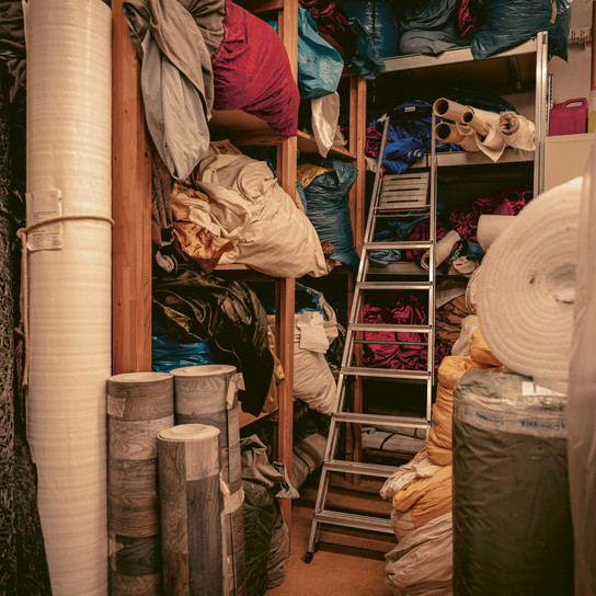 A cluttered storage room filled with various rolls and bundles of fabric in different colors, stacked on shelves and piled on the floor. A metal ladder is positioned in the center, amid the packed materials, indicating a busy textile space.