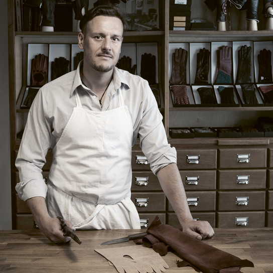 A craftsman in a white apron stands at a workbench holding scissors, with leather pieces spread out. Behind him are shelves displaying various gloves and a cabinet with multiple drawers.