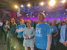 Eine Gruppe von Menschen, viele mit blauen "Chemnitz 2025 Volunteer"-Shirts, steht in einem Innenraum bei einer Veranstaltung. Sie lächeln und beobachten etwas aus dem Off, mit bunter Beleuchtung im Hintergrund.