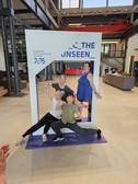 Vier Personen posieren spielerisch in einem großen Fotorahmen mit der Aufschrift "THE UNSEEN" in einer Veranstaltungshalle. Zwei Personen stehen, zwei kauern davor und lächeln. Das Display wirbt für Chemnitz als Kulturhauptstadt Europas 2025.