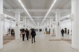 Eine geräumige, gut beleuchtete Industriehalle mit weißen Säulen und hohen Decken, in der die Menschen verstreut sind, einige unterhalten sich, andere gehen spazieren oder betrachten die Ausstellungen an den Wänden.