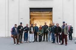 Eine Gruppe von elf Erwachsenen steht Seite an Seite vor einem großen, offenen Industrietor und posiert für ein Foto. Die meisten tragen Jacken und Freizeitkleidung. Der Hintergrund ist eine schlichte, helle Wand.