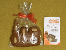 A gingerbread with almonds in the shape of a hand, wrapped in transparent plastic with a red bow, next to a leaflet with the inscription "Aachener Printen" on a yellow background.