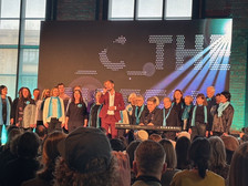 Ein Chor von Erwachsenen, von denen viele türkisfarbene Schals tragen, singt auf der Bühne vor einem Publikum. Ein Mann in einem kastanienbraunen Anzug steht in der Mitte. Im Hintergrund ist ein großer, beleuchteter Text zu sehen.