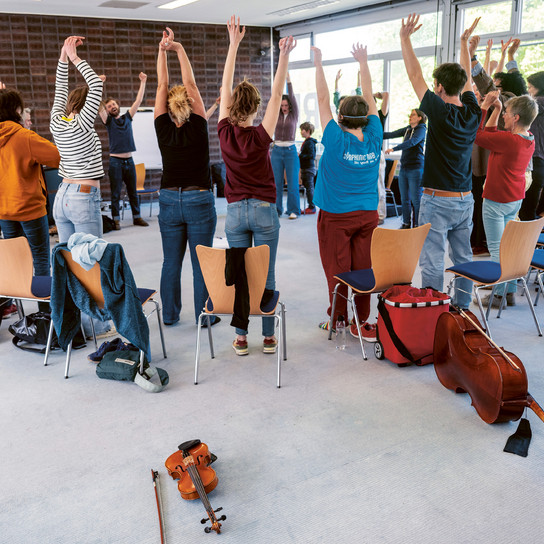 In einem hellen Raum mit großen Fenstern steht eine Gruppe von Menschen mit erhobenen Armen im Kreis. Stühle und Musikinstrumente, darunter eine Geige und ein Cello, sind im Raum verteilt.