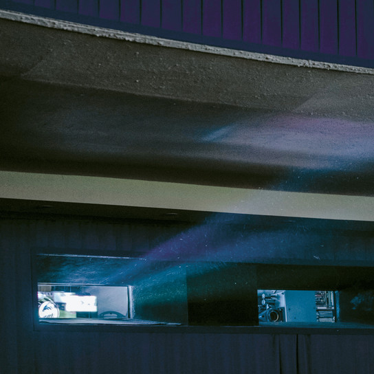 Ein Projektorstrahl fällt durch ein kleines Fenster in einen schwach beleuchteten Raum und verbreitet blaues und violettes Licht. Der Innenraum zeichnet sich durch niedrige Decken und dunkle Wände aus, was eine stimmungsvolle Atmosphäre schafft.