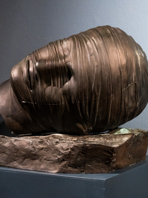 Eine dunkle Bronzeskulptur eines menschlichen Kopfes ist fest mit dünnen Bändern umwickelt und liegt seitlich auf einem Steinsockel vor einem neutralen Hintergrund. Das Gesicht ist teilweise von den Bändern verdeckt.