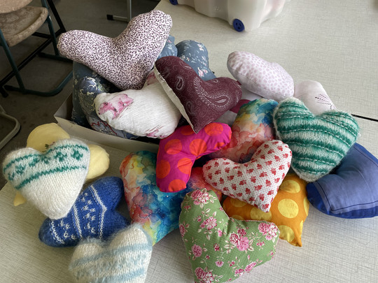 A collection of handmade heart-shaped cushions with various colourful patterns and fabrics, including knitted, floral, polka dot and patchwork designs, arranged on a table and in a box.