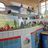 Eine bunte Auslage mit hölzernen Tier- und Baumfiguren ist auf einer gekachelten Theke bei einer Indoor-Veranstaltung aufgebaut. Auf einer oberen Ebene sind Menschen zu sehen, und Banner und Schilder schmücken den Hintergrund.
