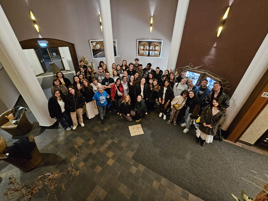 A large group of people pose for a photo in the lobby of a building with tiled floors, white columns and wall decorations. Most of them are smiling and looking at the camera, some are standing, others are sitting in front.
