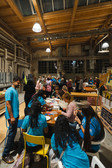 Eine große Gruppe von Menschen, viele in blauen Hemden, versammelt sich drinnen um Tische und arbeitet unter einer hohen Holzdecke in einem gut beleuchteten, industriell anmutenden Raum an Handwerks- und Kunstprojekten.