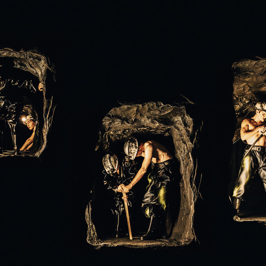 Drei kleine, schwach beleuchtete Abschnitte eines Bergwerks zeigen Figuren von Bergleuten mit Helmen und Arbeitskleidung, die vor einem dunklen Hintergrund unter Tage graben und schuften.