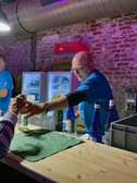 Ein Mann in einem blauen Hemd steht hinter einer Bar und reicht einem Kunden ein Getränk in Flaschen. Die Bar hat Glasflaschen, ein grünes Tuch und Kühlschränke mit Getränken im Hintergrund vor einer freiliegenden Ziegelwand.