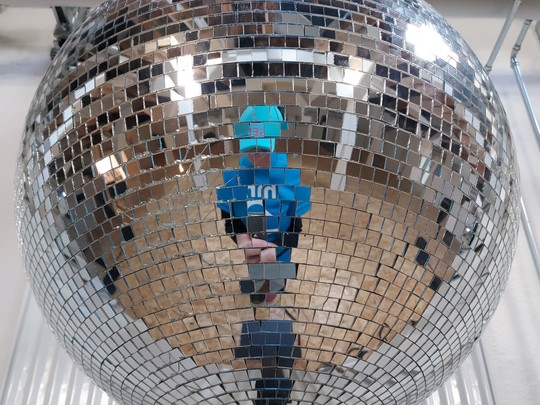 A person in a blue shirt and hat is reflected in a large, shiny disco ball made of mirrored tiles; a wooden floor and pipes can be seen in the background.