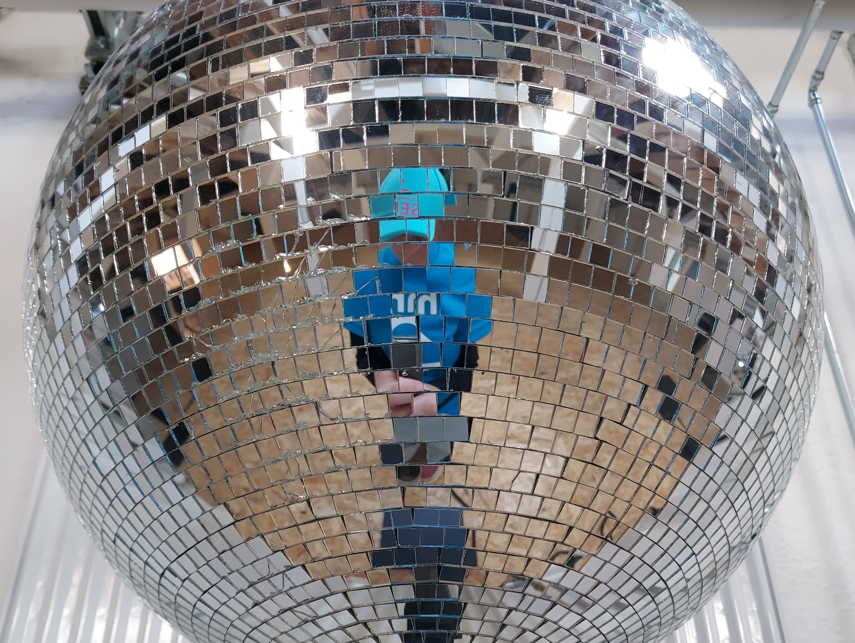 A person in a blue shirt and hat is reflected in a large, shiny disco ball made of mirrored tiles; a wooden floor and pipes can be seen in the background.