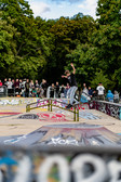 A skateboarder balances on a rail in a graffiti-covered skate park, surrounded by a crowd of onlookers and lush green trees in the background.