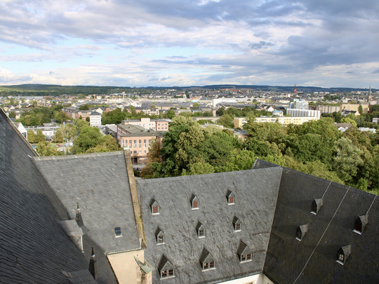 Eine Stadtansicht von oben zeigt schiefergedeckte Dächer mit dreieckigen Fenstern, grüne Bäume und eine weitläufige Stadt, die sich unter einem teilweise bewölkten Himmel in die Ferne erstreckt.