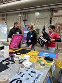 Drei Personen stehen an einem Basteltisch mit Farbe und Schablonen und bedrucken Tragetaschen in einer industriellen Innenumgebung. Auf dem Tisch sind Rollen mit Klebeband, Farbe und Kunstmaterialien ausgebreitet.