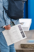 Eine hellblau gekleidete Person hält ein gefaltetes Papier mit dem fettgedruckten Titel "SICHT BETON" in der Hand und trägt eine dunkelblaue Tasche über der Schulter.
