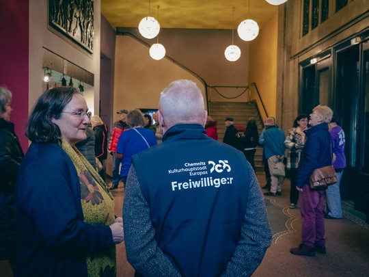 A group of people are standing in the entrance hall of a building and chatting. One person is wearing a waistcoat labelled "Volunteer". The atmosphere is relaxed; others are gathered or walking towards a staircase in the background. Spherical lights hang from the ceiling.
