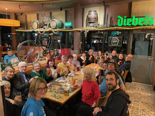 Eine große Gruppe von Menschen sitzt um zwei Tische in einem gemütlichen Restaurant, lächelt und erhebt die Gläser. Im Hintergrund sind dekorative Fässer, ein Fahrrad und Diebels-Bierschilder zu sehen. Alle sehen fröhlich und engagiert aus.