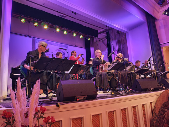 A band of musicians and singers perform on a stage with microphones and music stands, illuminated by colourful stage lights in a large interior with purple lighting.