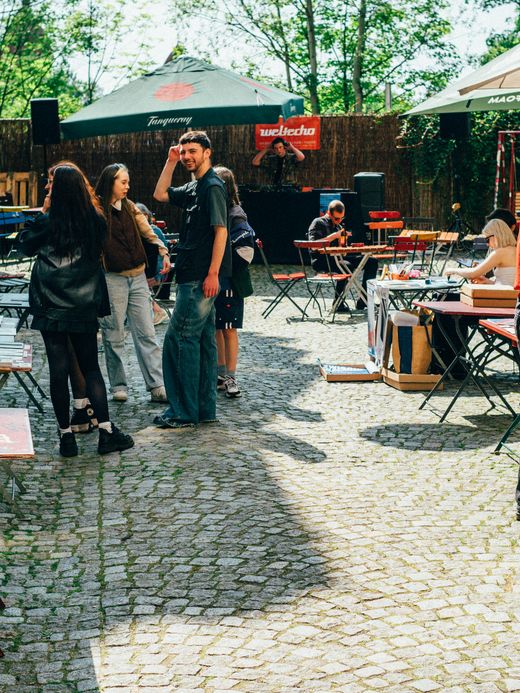 Menschen haben sich auf einer Kunstmesse oder einem Markt in einer Kopfsteinpflasterstraße versammelt. An den Tischen werden verschiedene Kunstwerke und Bücher ausgestellt. Sie stehen unter großen Sonnenschirmen und sind von Grün umgeben. Eine Person in einem roten Hemd betrachtet die Gegenstände auf einem Tisch genau.