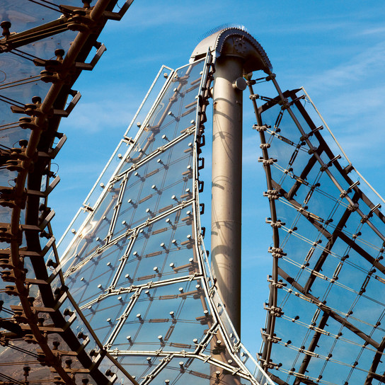 Eine Nahaufnahme einer modernen, gebogenen Glasstruktur vor blauem Himmel. Das Design besteht aus einer Reihe von Metall- und Glasplatten, die mit einer zentralen Säule verbunden sind und so ein künstlerisches und architektonisches Erscheinungsbild erzeugen.