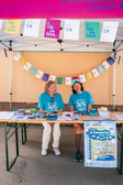 Zwei Freiwillige in blauen "Chemnitz 2025"-Shirts sitzen an einem mit Bannern und Flyern geschmückten Informationsstand unter einem rosa Zelt. Auf dem Tisch liegen Broschüren, Bücher und Kleinigkeiten.