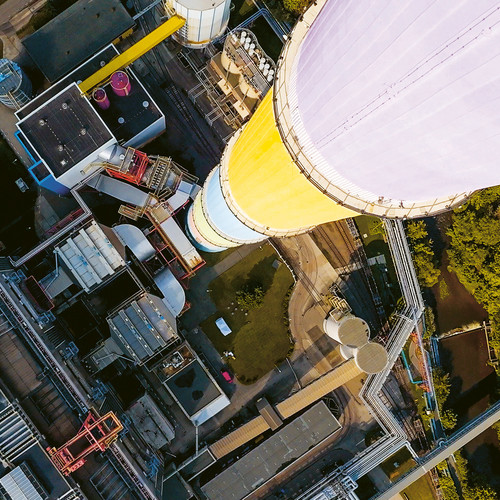 Luftaufnahme eines Industriekomplexes mit einem großen, farbenfrohen zylindrischen Turm. Der Turm besteht aus Abschnitten, die in Weiß, Gelb, Blau und Lila gestrichen sind. Die Umgebung umfasst Gebäude, Wege und Grünflächen.