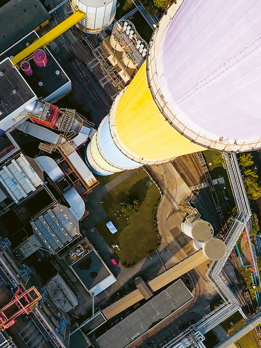 Luftaufnahme eines Industriekomplexes mit einem großen, farbenfrohen zylindrischen Turm. Der Turm besteht aus Abschnitten, die in Weiß, Gelb, Blau und Lila gestrichen sind. Die Umgebung umfasst Gebäude, Wege und Grünflächen.