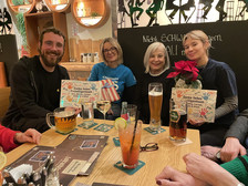 Vier Personen sitzen um einen Holztisch in einer gemütlichen Bar und lächeln in die Kamera. Drei halten Danksagungskarten und einen Weihnachtsstern. Verschiedene Getränke und Untersetzer stehen auf dem Tisch. Hinter ihnen steht eine Tafel mit deutscher Schrift.