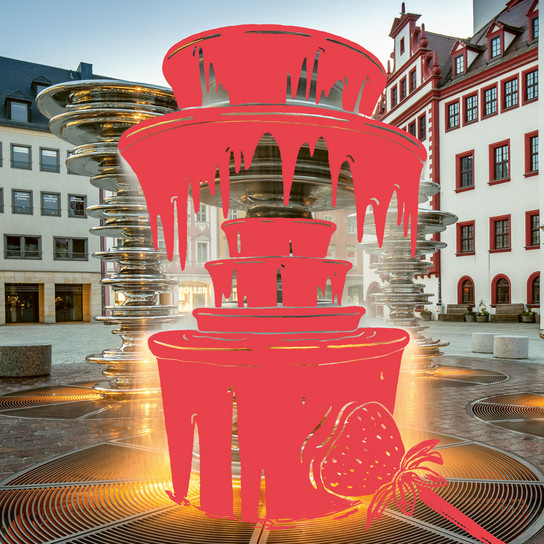 Ein digital bearbeitetes Bild zeigt einen Stadtbrunnen mit einer Grafiküberlagerung, die einem Schokoladenbrunnen mit roter tropfender Schokolade und einer Erdbeere ähnelt. Im Hintergrund sind historische Gebäude auf einem Kopfsteinpflasterplatz zu sehen.