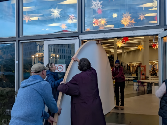 Vier Personen arbeiten zusammen, um ein großes, gekrümmtes Objekt durch doppelte Glastüren in ein hell erleuchtetes Gebäude zu bringen, das mit bunten sternförmigen Lichtern und einem Weihnachtsbaum im Inneren geschmückt ist.