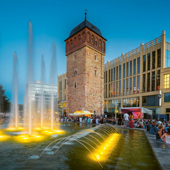 Eine geschäftige Abendszene mit Menschen, die sich um einen großen, beleuchteten Brunnen versammelt haben. In der Nähe steht ein historischer Backsteinturm, neben einem modernen Glasgebäude. Der Himmel ist klar und trägt zur lebhaften Atmosphäre bei.