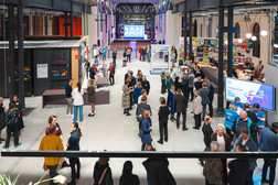 Eine große Indoor-Veranstaltung mit vielen Menschen, die sich mischen, unterhalten und Stände erkunden. Helle Lichter erhellen die geräumige Halle mit ihren hohen Decken und einer Bühne mit einer Leinwand am anderen Ende. Einige Leute stehen in Gruppen oder gehen umher.