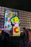 Auf einem nächtlichen Backsteingebäude ist eine farbenfrohe digitale Kunstprojektion zu sehen, die eine große kohlartige Figur, lebhafte Muster und dekorative Motive zeigt.