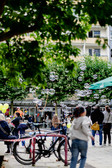 People stroll through a lively outdoor area where soap bubbles float in the air, bicycles are parked in the foreground and trees and balloons add colour and vibrancy to the scene.