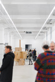 Eine Gruppe von Menschen steht und unterhält sich in einer hellen, modernen Kunstgalerie mit hohen Decken. Im Hintergrund sind Pappkartons und Skulpturen ausgestellt. Die Szene wirkt ungezwungen und informell.