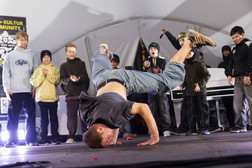A break dancer balances on one hand with his legs outstretched in the air and performs on stage while a group of children watch and cheer in the background. The scene is lively and full of energy.