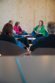 Five people sit in a semi-circle and discuss in a modern room with light-coloured wooden walls. A woman in a green jacket seems to be leading the conversation.