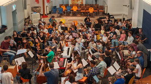 Ein großes Orchester probt in einem hell erleuchteten Raum. Die Musiker spielen verschiedene Instrumente, darunter Streicher, Holzbläser, Blechbläser und Keyboards. Bunte Stühle und Notenständer füllen den lebendigen Raum.