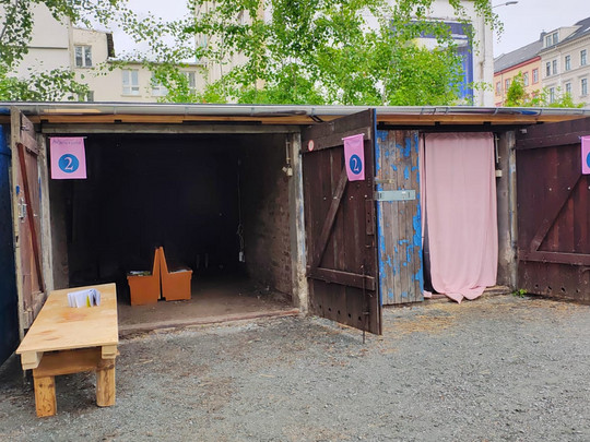 Three wooden stands with open doors are labelled with the numbers 1, 2 and 3. Stand 1 has a table with papers in front of it, stand 2 is empty and stand 3 has a pink curtain. The area is covered with gravel flooring and buildings in the background.